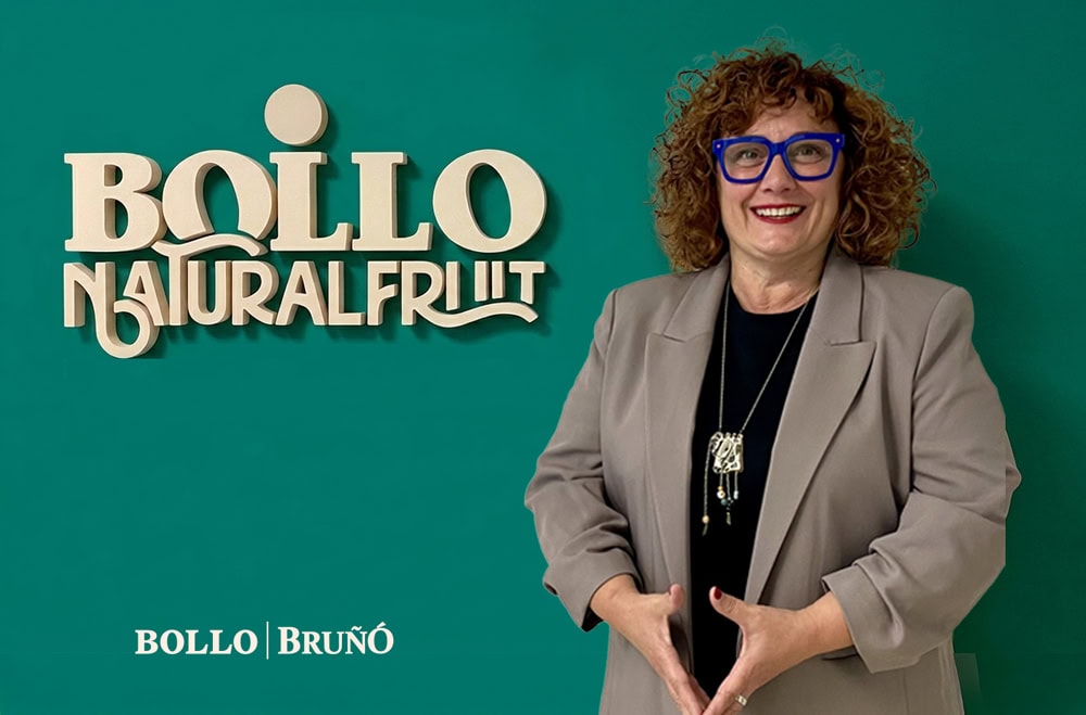 Smiling woman with curly hair and blue glasses poses beside a teal wall with a 'BOLLO Natural Fruit' sign and branding in the corner.