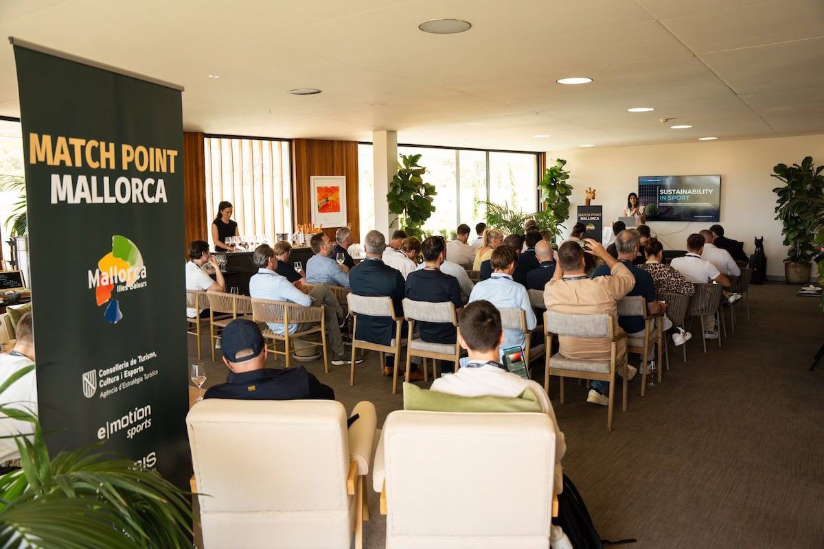 Conference audience seated in a bright room with a tall green banner reading 'MATCH POINT MALLORCA' at left and a speaker at the front podium on screens on the right.