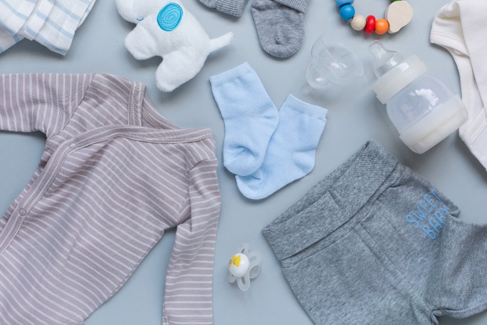Flat lay of baby clothes and accessories: striped onesie, gray shorts with 'Sweet Baby', blue socks, bottle, pacifier, plush toy, and teething beads on a blue background.