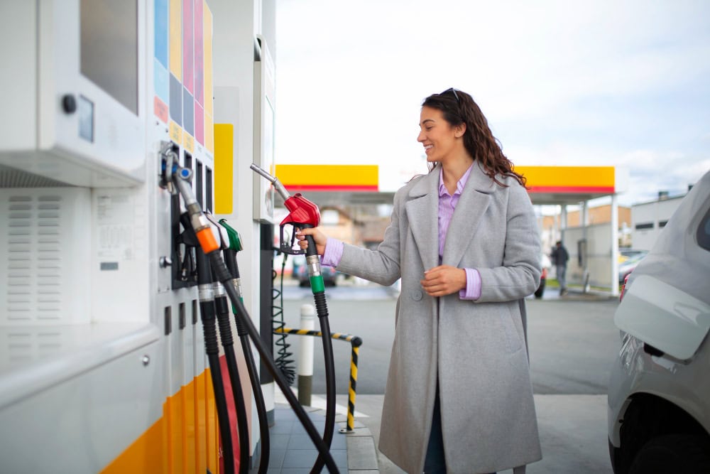 Woman in a gray coat pumps gas at a fuel station beside a parked car, smiling.
