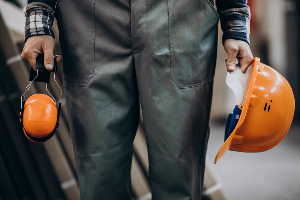 Worker in gray-green coveralls holds an orange hard hat in one hand and orange earmuffs in the other, ready for construction work.
