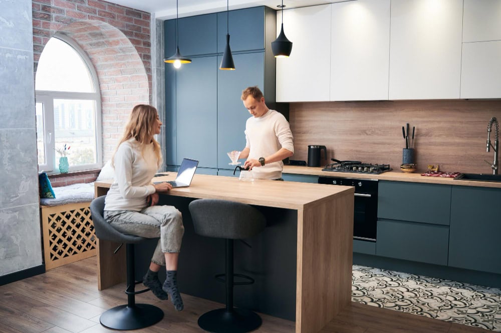 Woman sits at a wooden kitchen island with a laptop while a man pours coffee beside her in a modern blue-gray kitchen with pendant lights and brick wall.