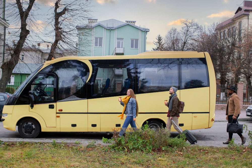 Three pedestrians walk beside a large yellow bus on a city street, each carrying a drink/cup homewardbound.