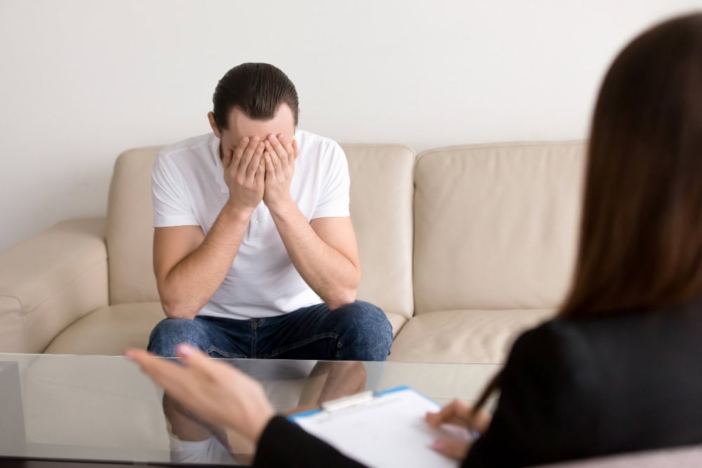 Distressed man in a white shirt sits on a beige sofa, covering his face with his hands during a therapy session.
