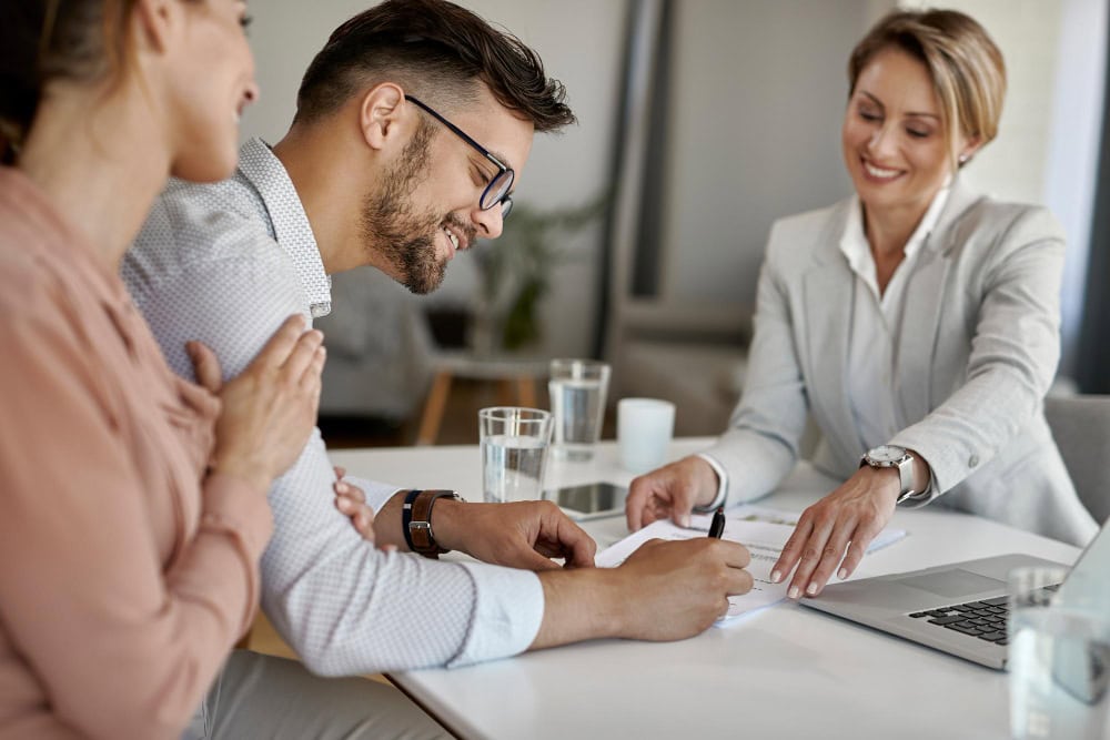 Business meeting: a man signs documents at a table while a woman in a light blazer guides him, with colleagues watching.