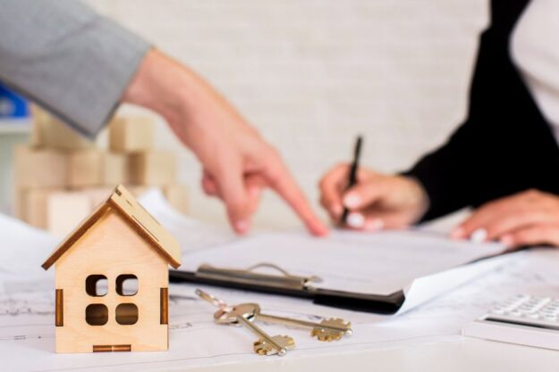 Wooden house model, keys, and documents on a desk during a real estate signing scene with a person writing and another pointing at papers