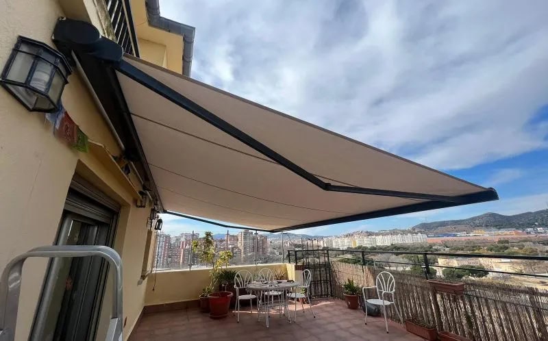A spacious balcony with a beige retractable awning extended, metal table and chairs, potted plants, and a city skyline in the distance.
