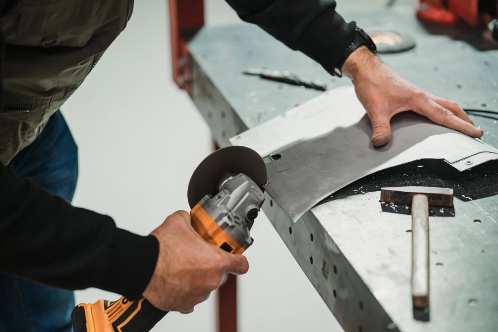 Worker using an angle grinder to cut a metal sheet on a workbench, with a hammer nearby and tools in the background