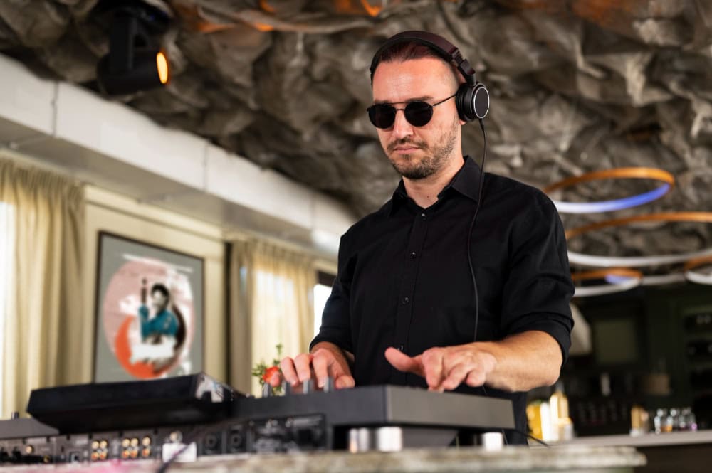 DJ wearing sunglasses and headphones, mixing on a keyboard-style controller at a bar/club setup