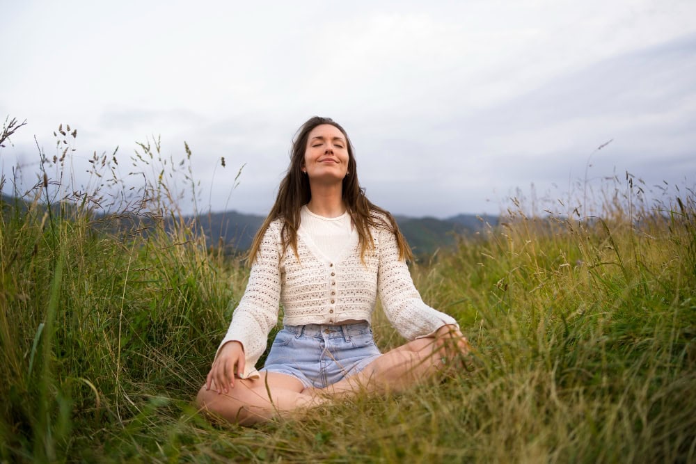 Woman sits cross-legged in tall grass, meditating with eyes closed in a meadow hillside.