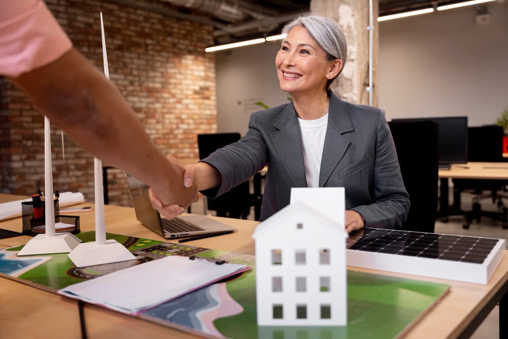 Businesswoman and client shake hands over a desk with a white model house and a solar panel beside them.