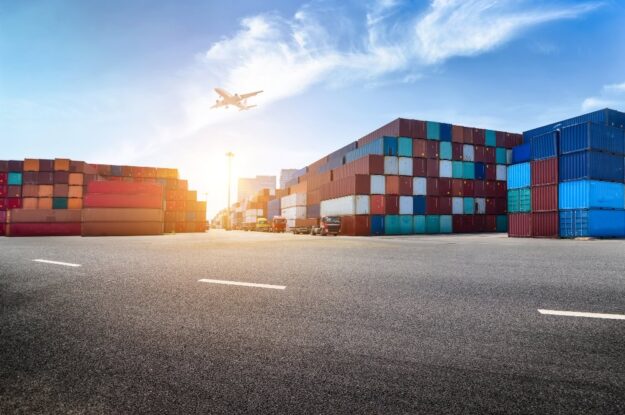 Large outdoor shipping yard with stacked colorful shipping containers, trucks in the distance, and an airplane flying low in a bright blue sky with sun glare on the horizon.