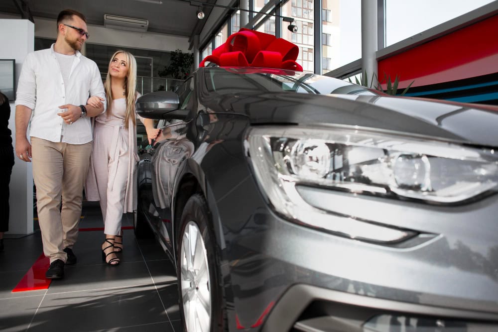 Couple walking beside a new gray car with a large red bow in a showroom.