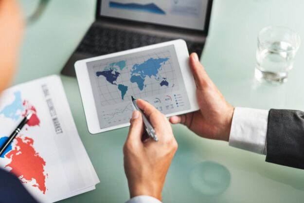 Businessperson holding a tablet showing a world map and charts, with a laptop, printed maps, and a glass of water on a glass desk.