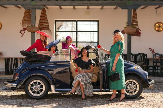 Five women in vintage dresses and hats pose with a dark blue convertible in a sunny courtyard.