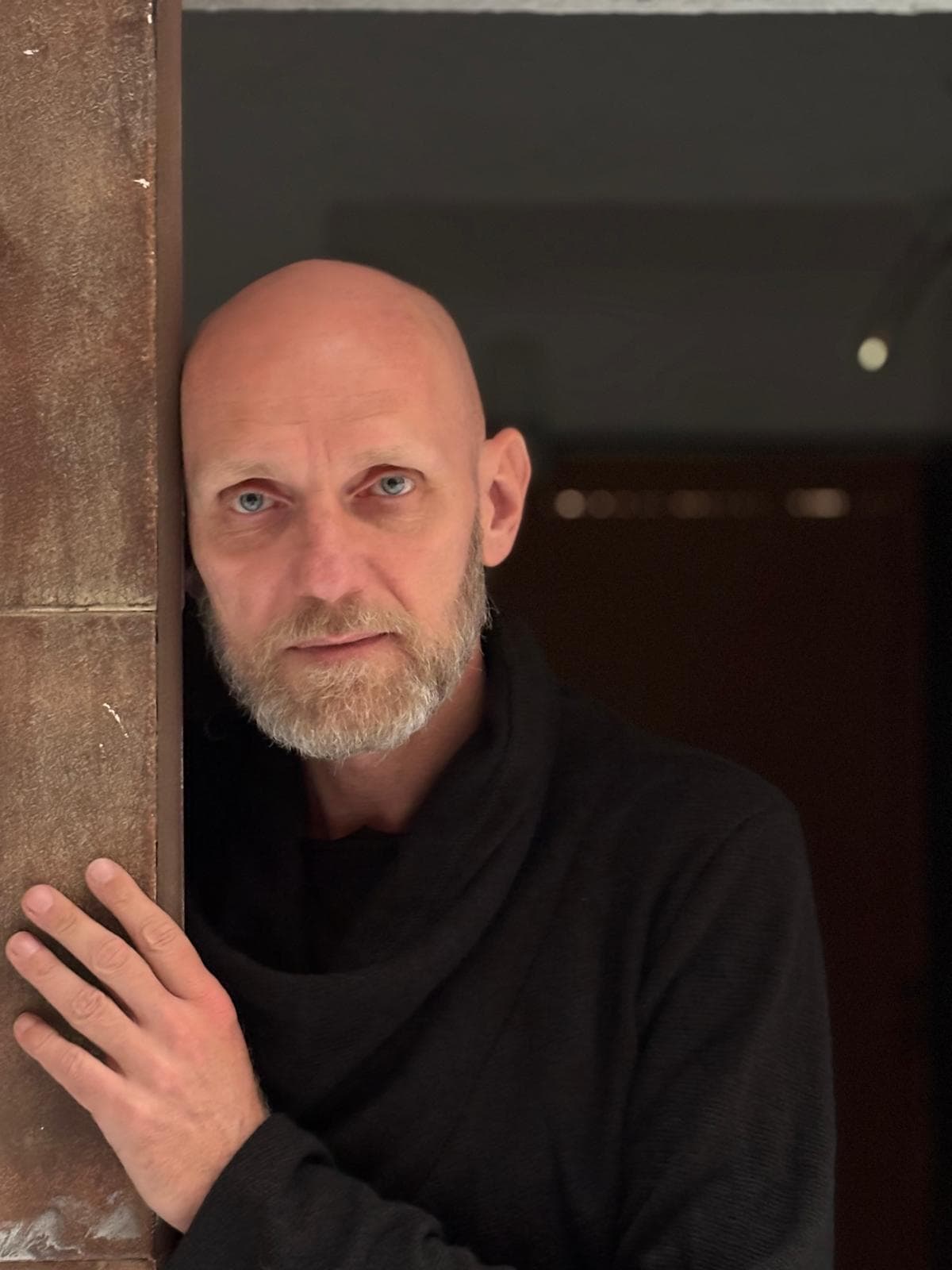 Bald man with a gray beard leaning against a wooden door frame, wearing a black sweater and looking at the camera.