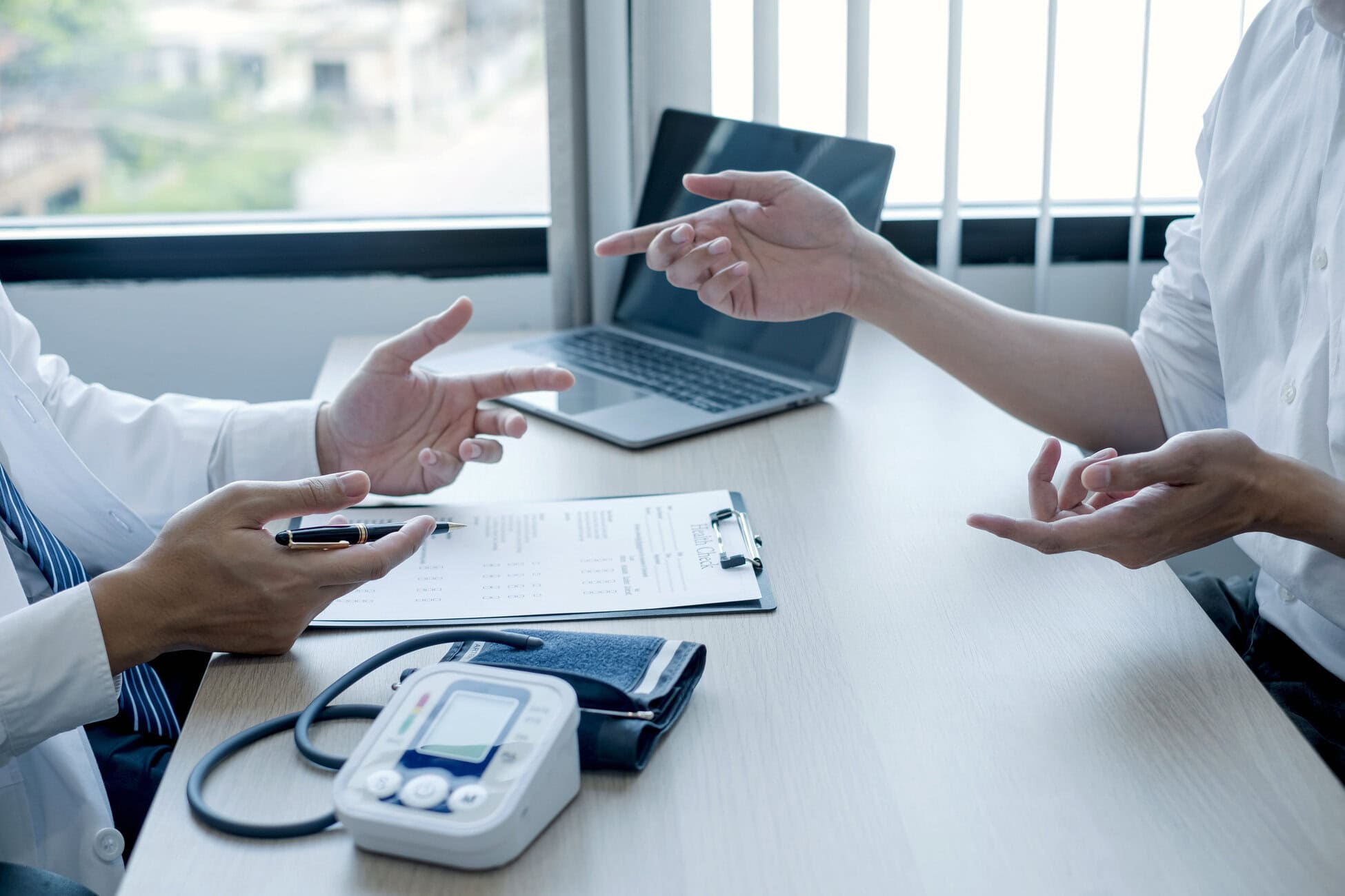 Medical professionals discuss a chart at a table with a laptop and medical device nearby in a bright office.