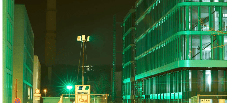 Nighttime street view of a modern glass building under construction, illuminated in green with construction barriers in the foreground.