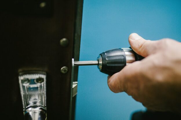Person uses a cordless drill to drive a screw into a door strike plate on a dark door, with a blue backdrop behind.