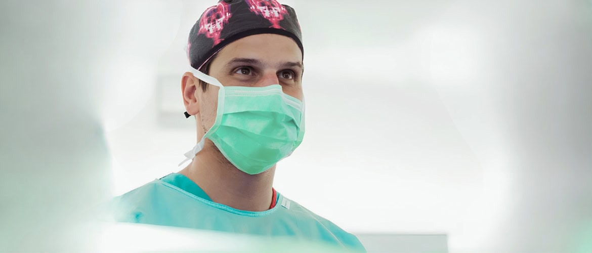 Surgeon wearing a green surgical mask and cap, looking upward in a bright clinical setting.