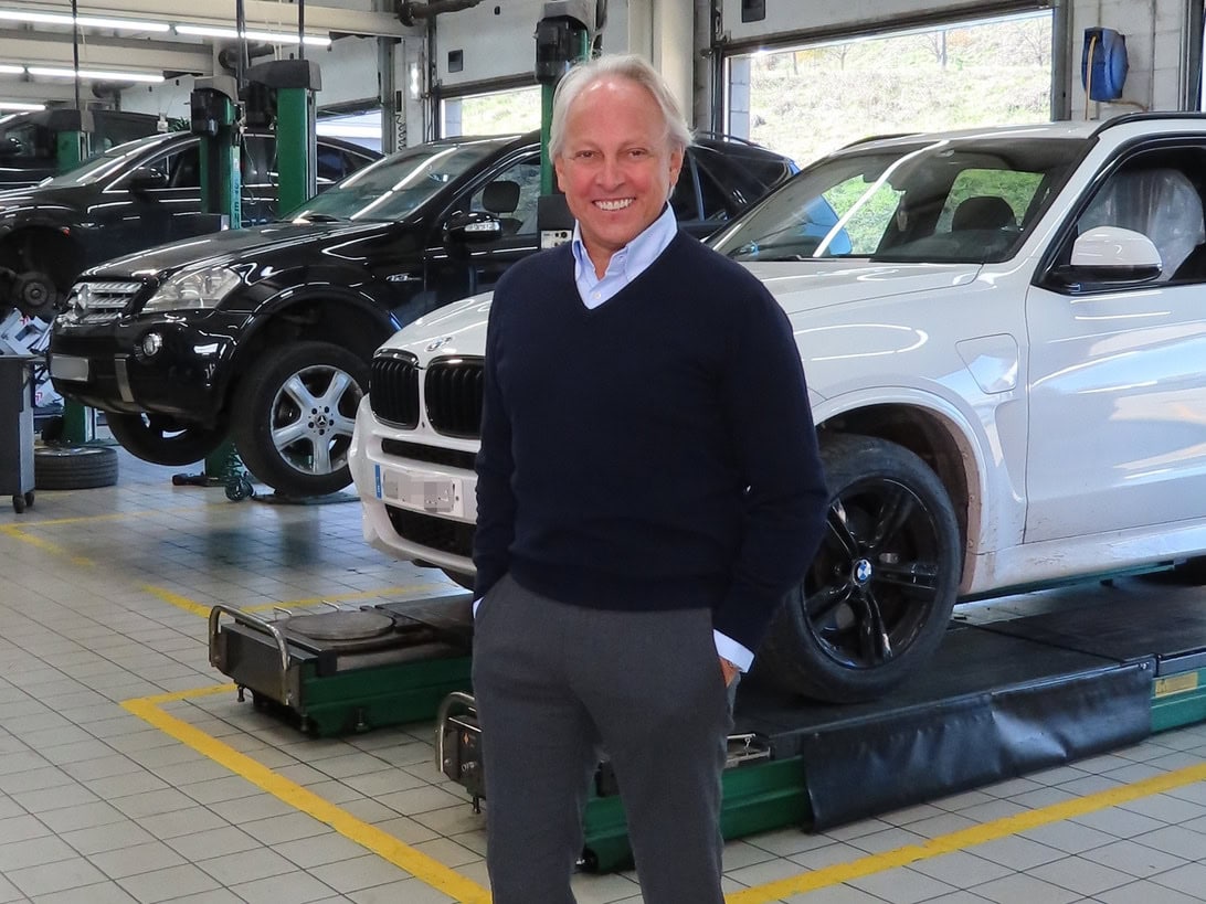 Smiling man in a navy sweater stands in an auto repair shop with a white SUV on a lift behind him.