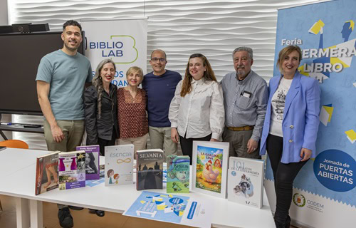 Eight people posing behind a table with displayed books at a library event, banners visible in the background.