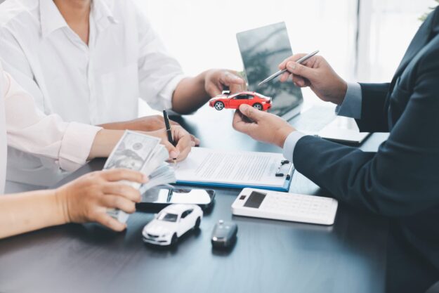 Business meeting: a person in a suit hands a red toy car to another person while reviewing documents on a desk with model cars and a calculator.
