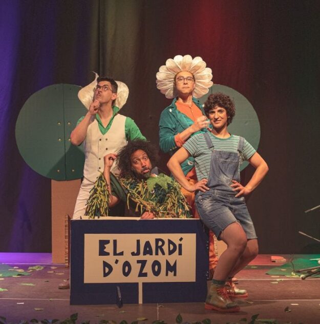 Four actors in bright, whimsical costumes perform on a theater stage; a prop box in front is labeled 'EL JARDÍ D'OZOM'.