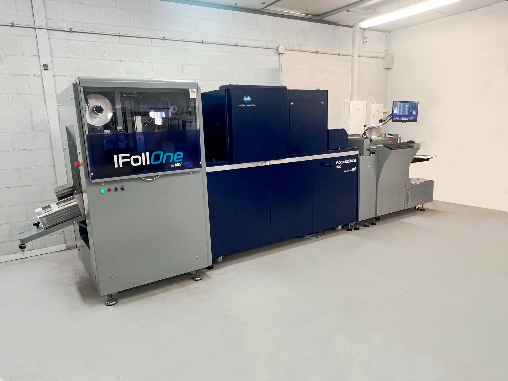 Industrial foil-stamping and finishing machine line (iFoilOne by MGI) in a clean white-walled room, with a conveyor on the left and a touchscreen monitor on the right.