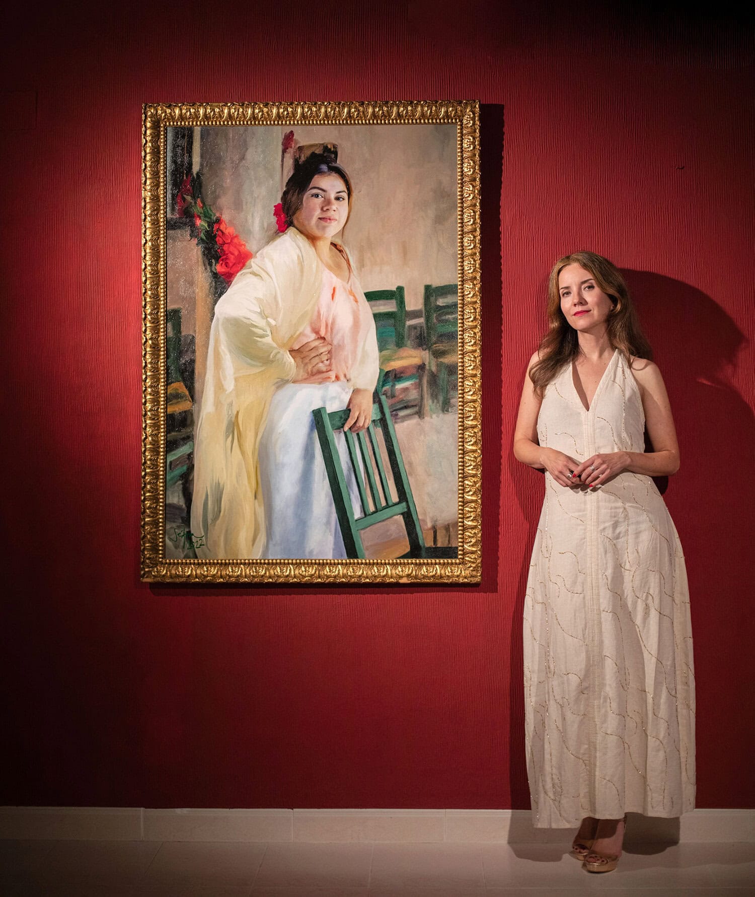 Woman in a white sleeveless dress standing beside a large framed painting on a red gallery wall.