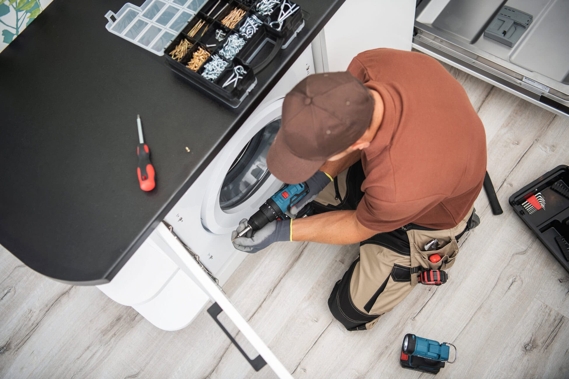 Overhead view of a technician kneeling by a front-loading washing machine, using a drill with a toolbox and tools nearby on a dark counter.