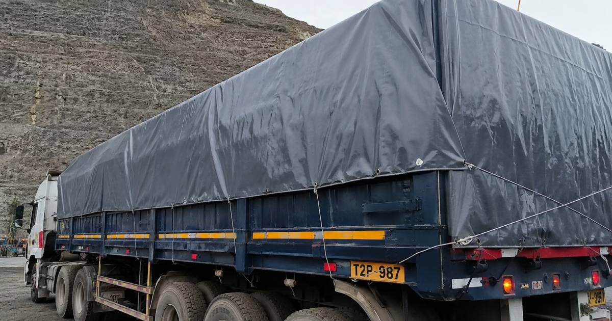 Blue flatbed truck with a large grey tarpaulin cover, parked near a rocky cliff; trailer shows yellow safety stripes and license plate T2P-987.