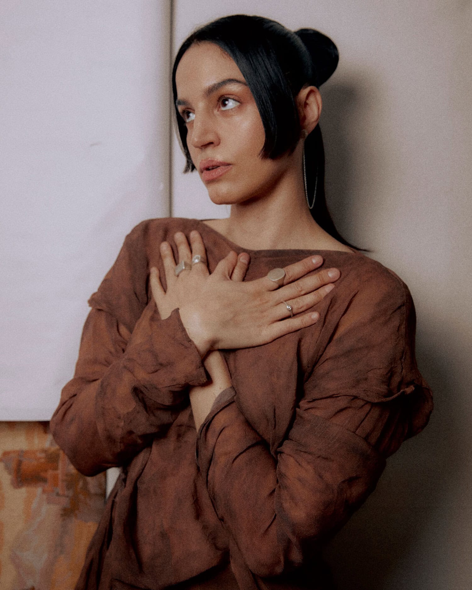 Woman in a brown, textured dress crosses her arms over her chest while leaning against a wall, looking to the left with a calm expression, wearing hoop earrings and rings on several fingers.
