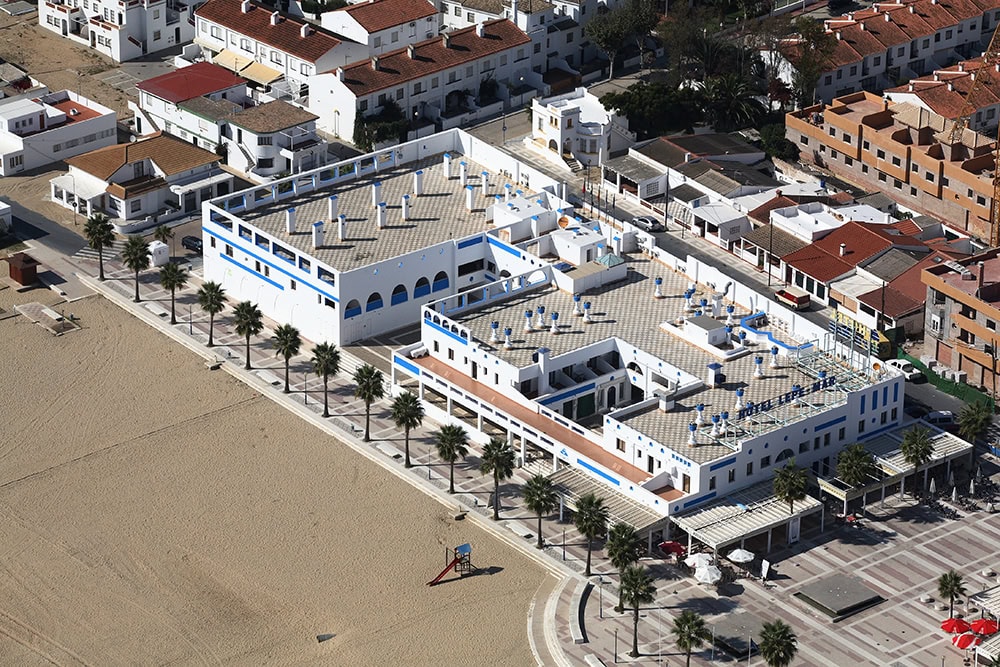Aerial view of a white seaside hotel complex with blue accents along a sandy beach and palm-lined promenade.