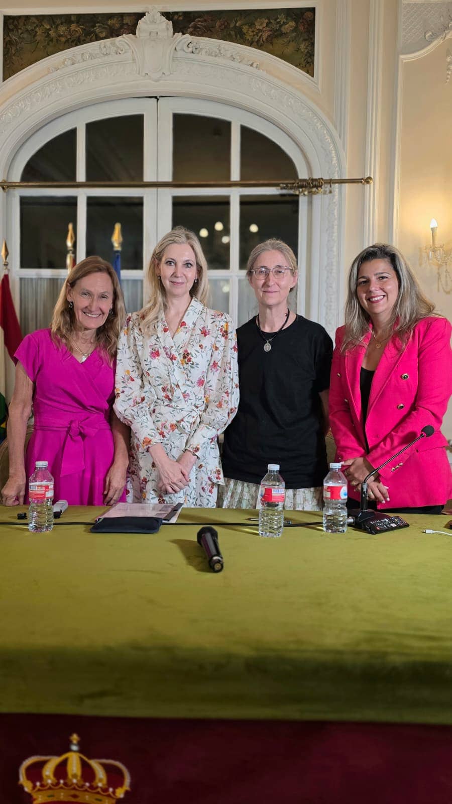 Four women stand together behind a table with microphones in an ornate room, posing for a group photo at a panel event.