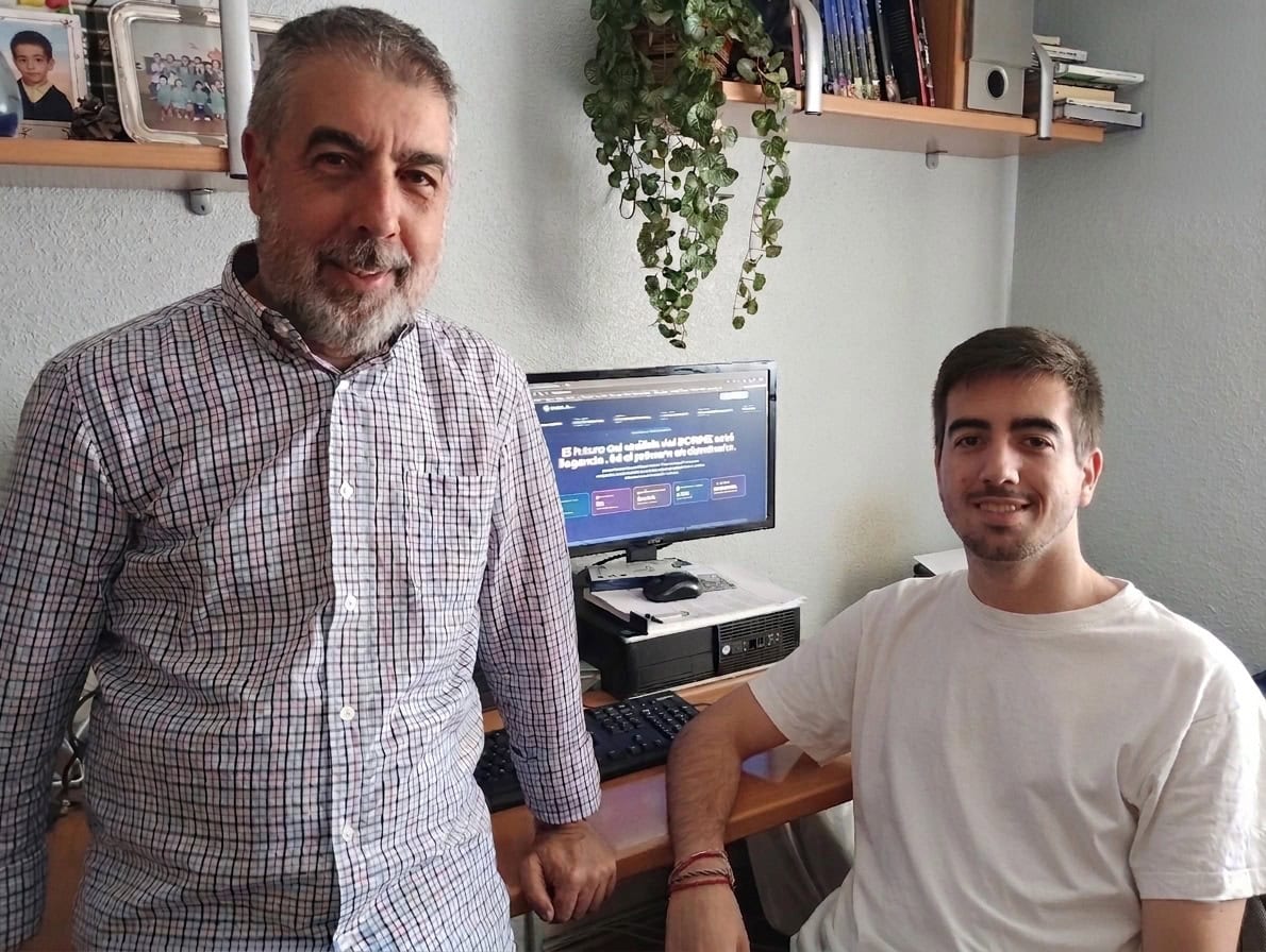 Two men in a home office: an older man in a checkered shirt stands by a desk while a younger man in a white shirt sits at the computer smiling.