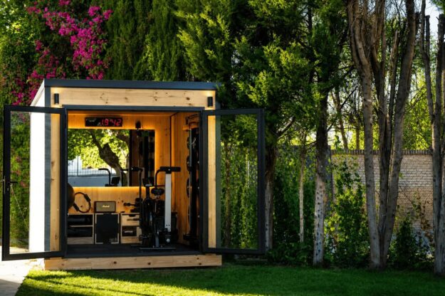 Small wooden shed converted into a home gym, with open glass doors and exercise equipment inside, set in a sunny backyard with trees nearby.