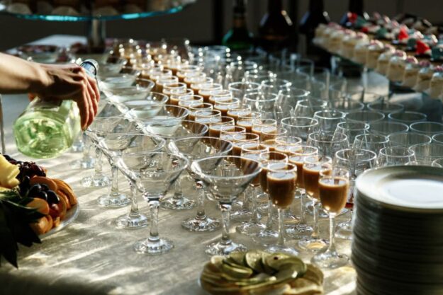 Catered table with rows of champagne flutes being poured, fruit platters visible to the side.