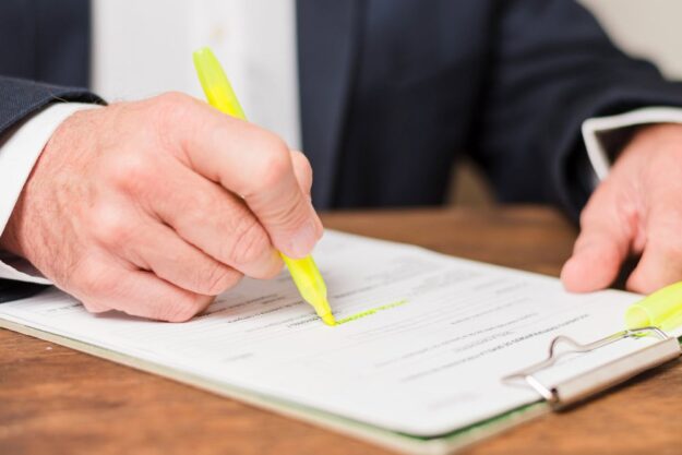 A person in a dark suit signs a document with a yellow highlighter on a clipboard.