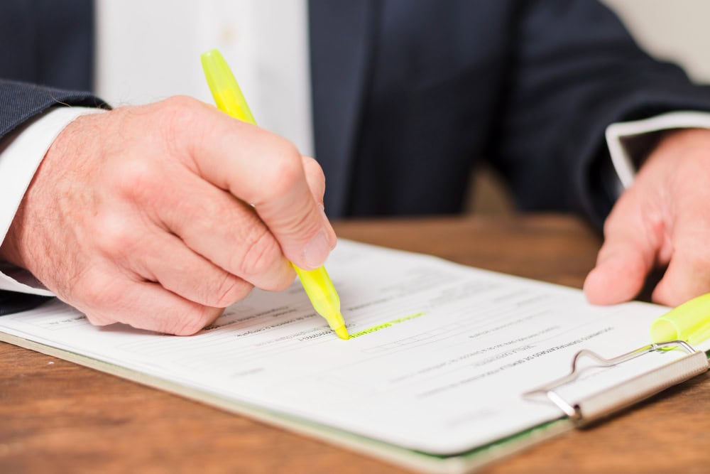 A person in a dark suit signs a document with a yellow highlighter on a clipboard.