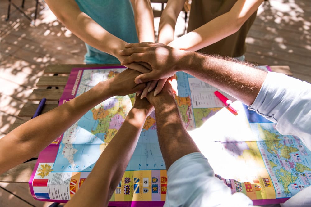 Group of people placing hands together over a world map on a table, symbolizing teamwork and travel planning.