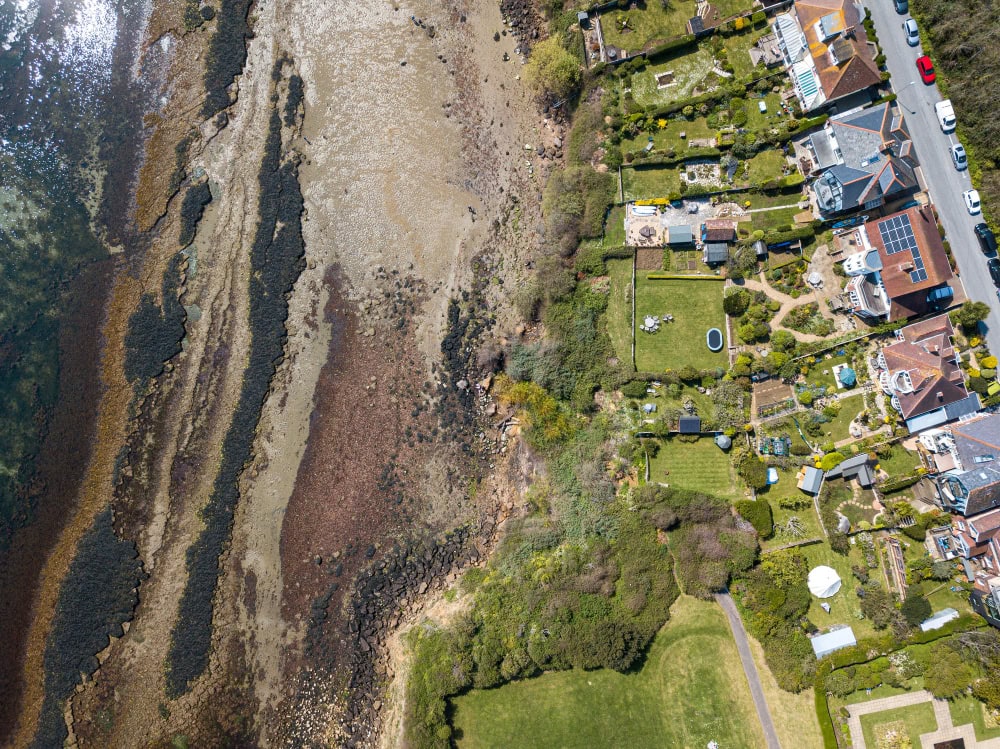 Aerial view of a rocky shoreline with water on the left and a tidy seaside neighborhood with lawns and houses on the right.