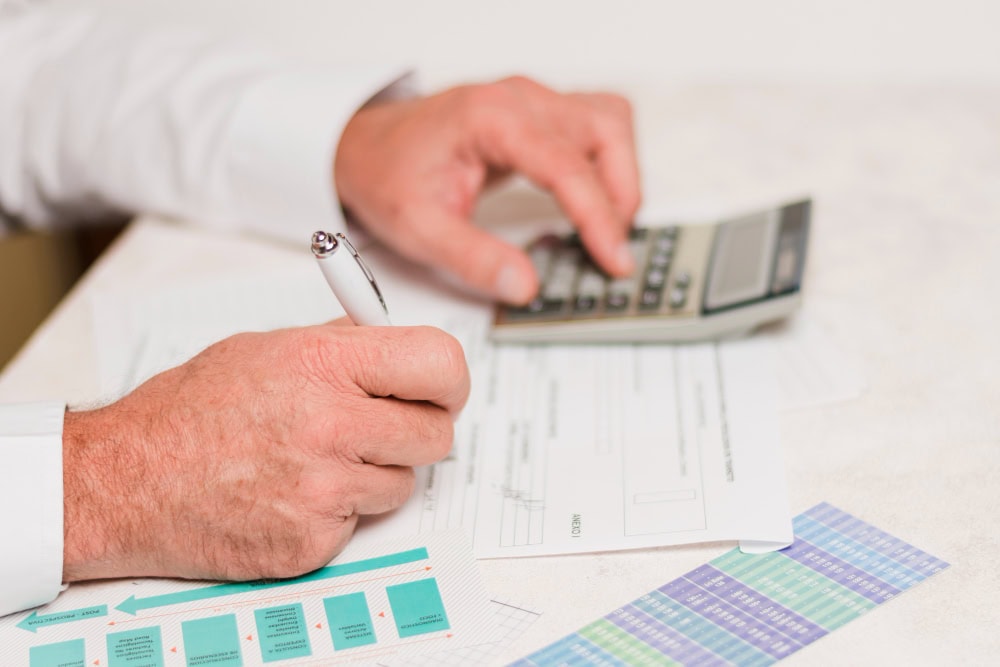 Hands write on forms with a pen while using a calculator; paperwork and color charts are on the desk.