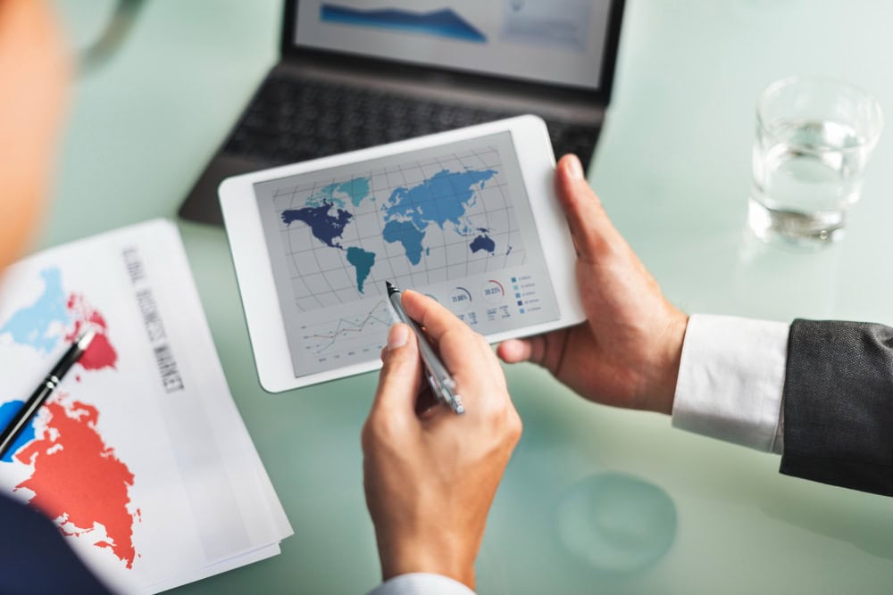 Person in business attire uses a tablet displaying a world map and charts during a meeting, with a pen and a printed color map on the desk and a laptop in the background.