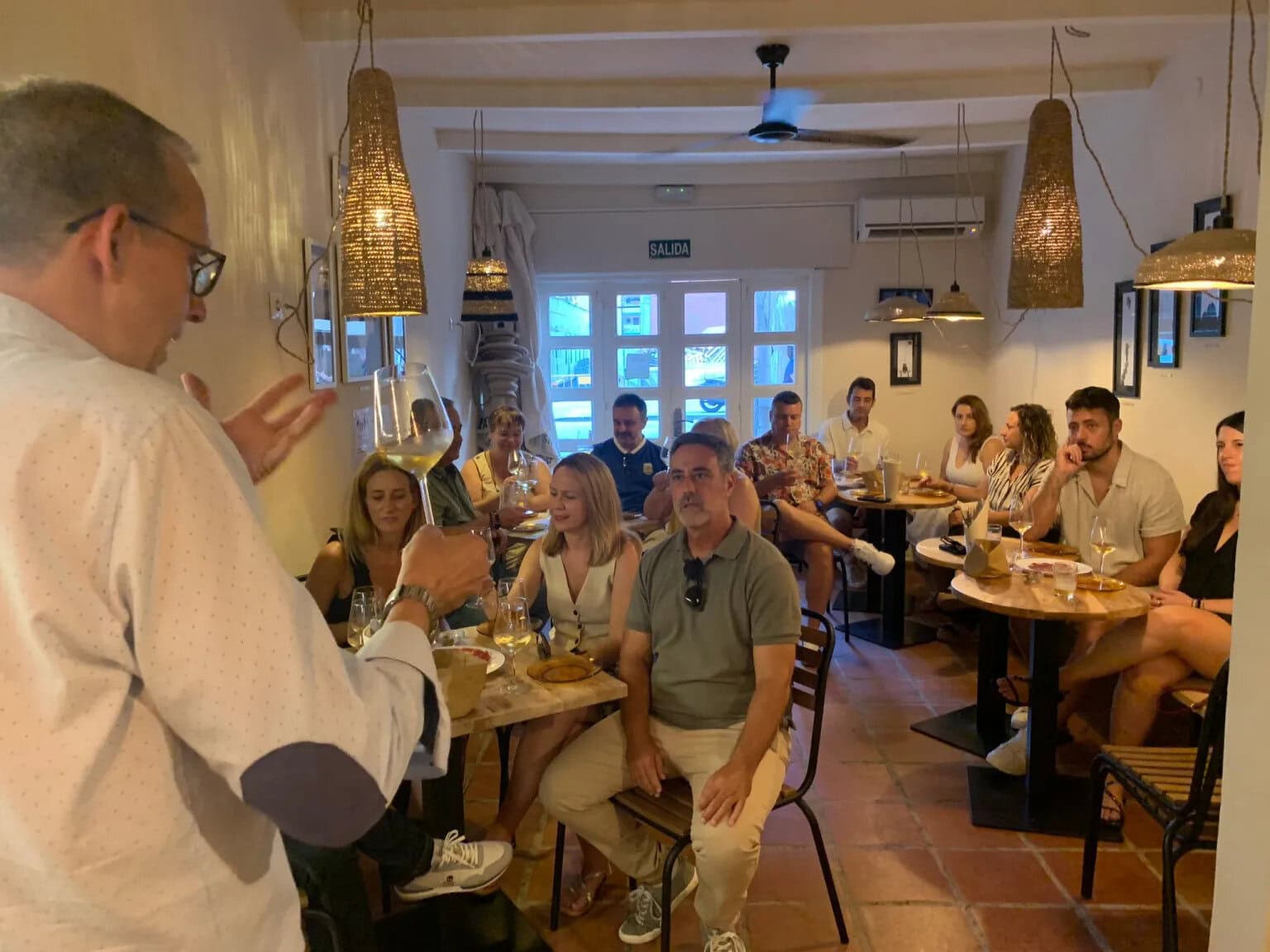 A man with a white shirt speaks to a seated group in a cozy restaurant, raising a large wine glass as the others listen around wooden tables with drinks and plates.