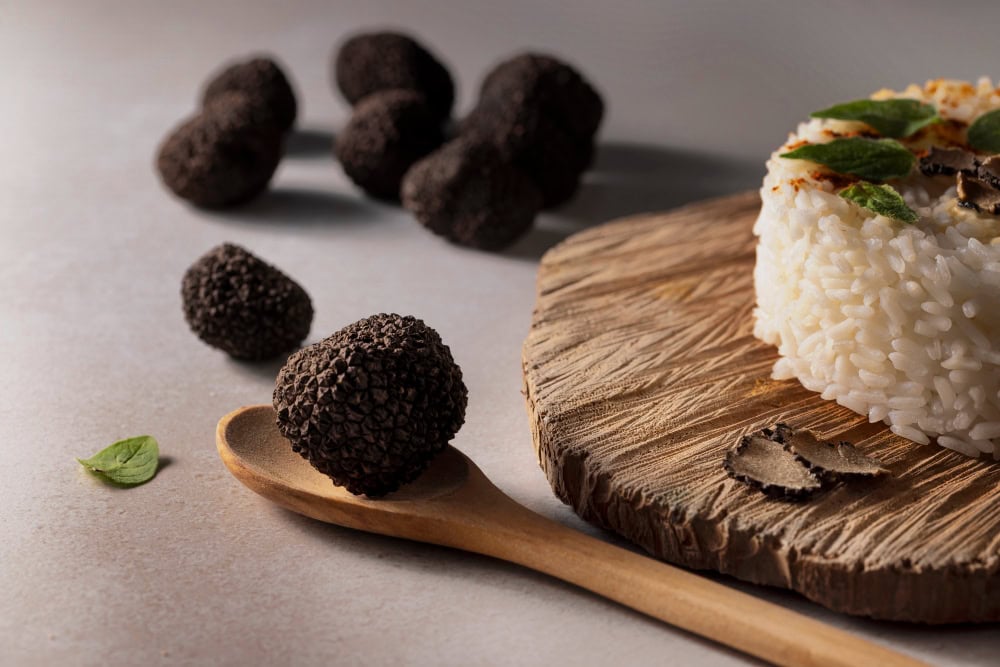Rice dish shaped on a rustic wooden board, surrounded by whole black truffles and a wooden spoon with a truffle on it.