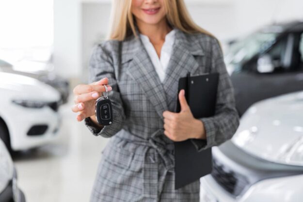 Woman in a gray plaid blazer extends a car key fob toward the camera in a showroom of parked cars.
