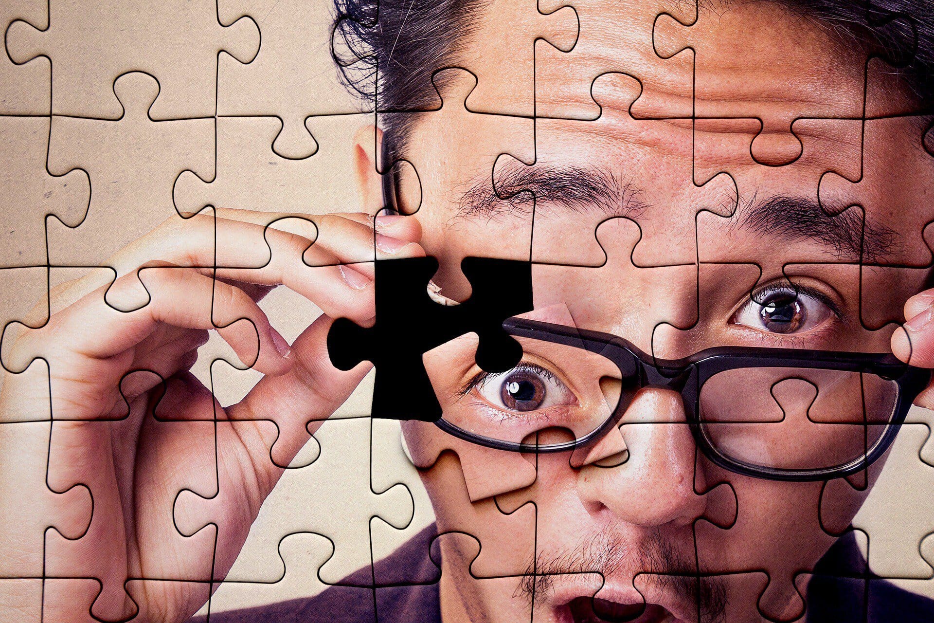 Close-up of a man with glasses, placing a puzzle piece near his eye while puzzle pieces cover his face.