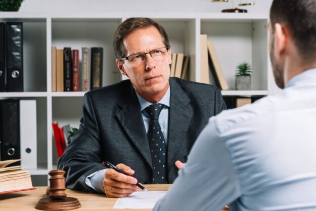Lawyer in a dark suit and glasses sits at a desk with a gavel, discussing documents with a client across the table.
