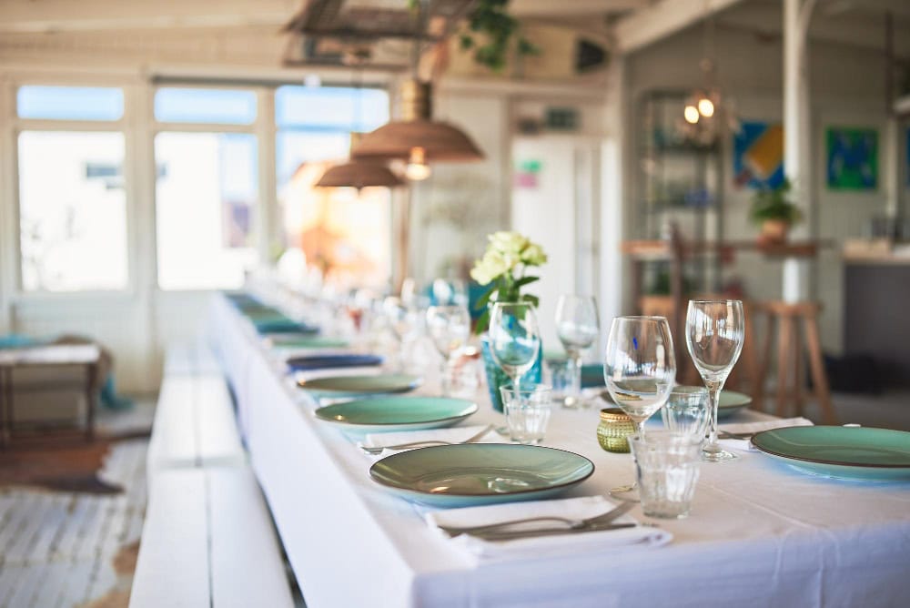 Long table set for a meal with green plates, glassware, and flowers in a bright, airy dining room, ready for guests.